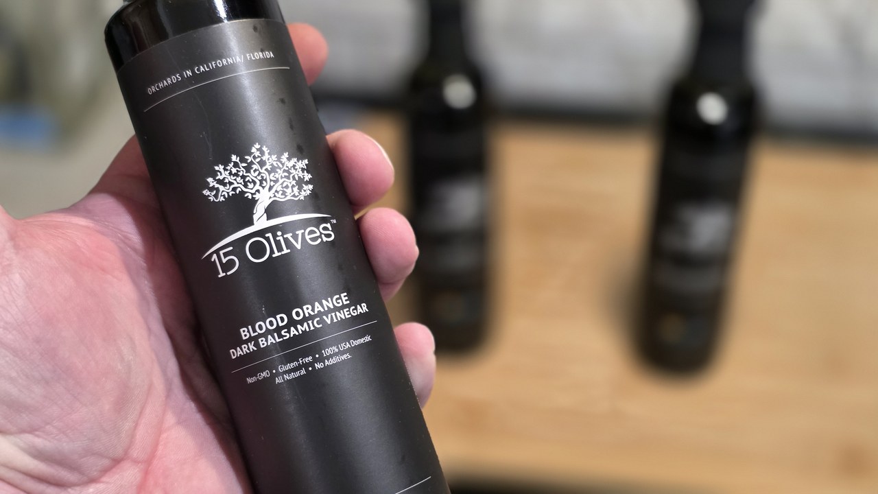 15 Olives Blood Orange Dark Balsamic Vinegar bottle held in hand, showing California and Florida orchards label, with two soft-focus 15 Olives bottles on a wooden surface in the background