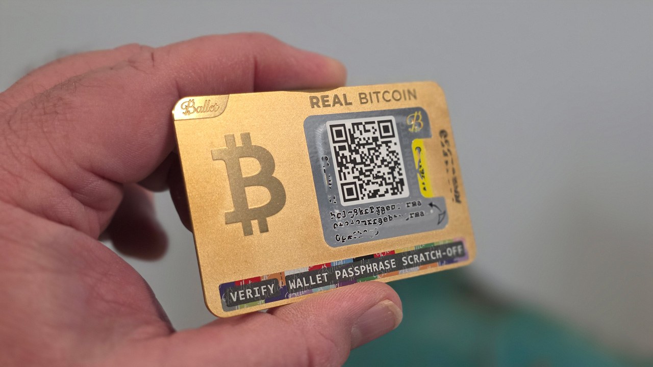 Gold-plated Ballet REAL Bitcoin cold storage wallet held in hand, showing front QR deposit address and lottery-style scratch-off passphrase strip
