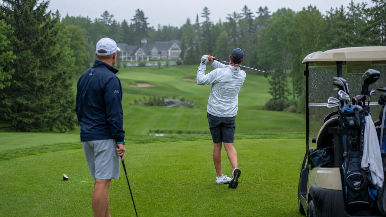 Two men golf in light rain on a tree-lined northern course, one finishing his swing on the tee box beside a cart loaded with golf bags