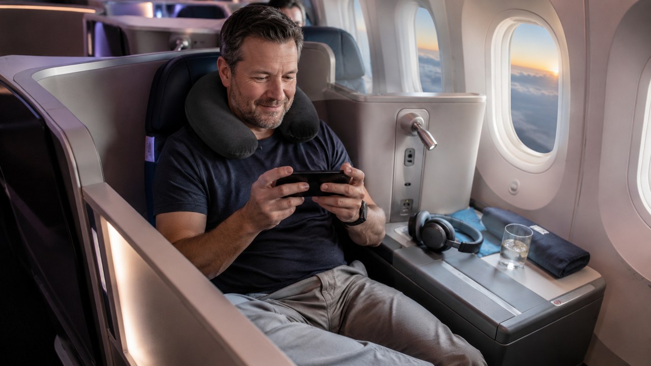 Man in a business-class seat wearing a travel neck pillow and playing a game on his phone during a long-haul flight, with noise-canceling headphones resting on the console