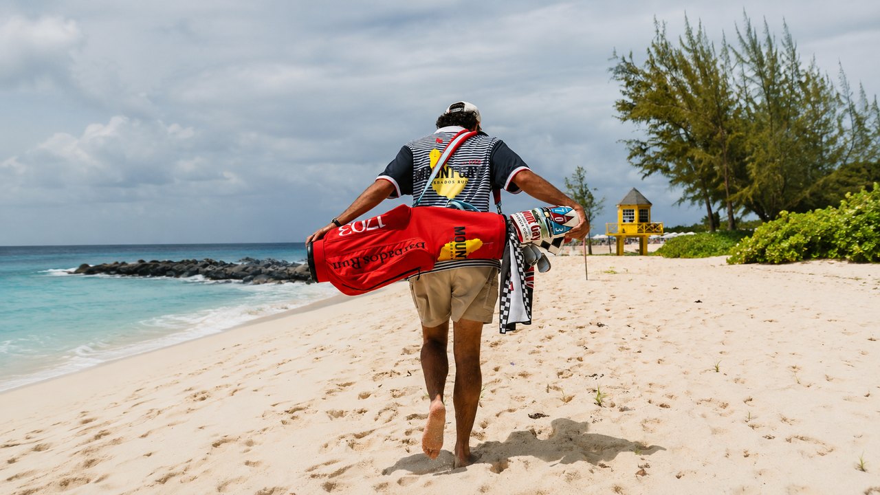 A golfer walks a Barbados beach carrying the red Mount Gay 1703 Devereux Golf bag and checkered towel