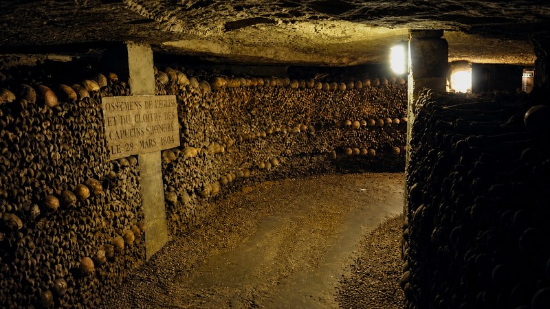 Long corridor in the Paris Catacombs lined with stacked bones and skulls, with a marble plaque visible on the left