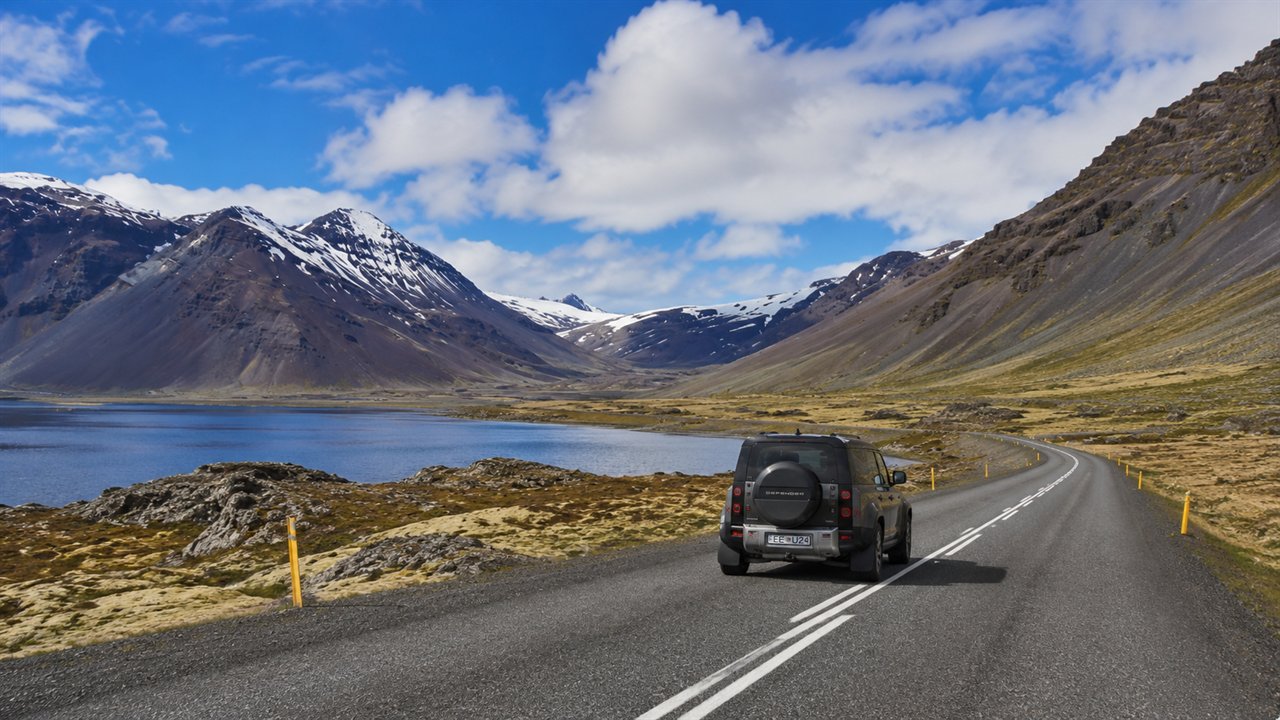 Land Rover Defender on a paved Iceland road beside a fjord and snow-capped mountains