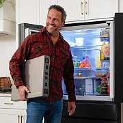 A man carries the slim 4Patriots Blackout Buddy emergency power supply past an open, fully stocked refrigerator in a bright kitchen