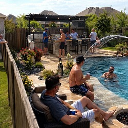 Guys hanging out around a backyard pool with a fire pit, outdoor kitchen and a pergola strung with lights