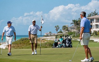 Four men golfing on an Emerald Coast course above the Gulf, with a cart and beach houses behind them
